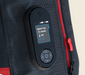 A close-up of a black handheld electronic device with a small color display and directional pad, attached to or placed on a dark textured bag with red zipper accents.